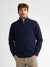 A man wearing a navy blue quarter-zip knit sweater and beige trousers, standing against a plain light gray studio background.