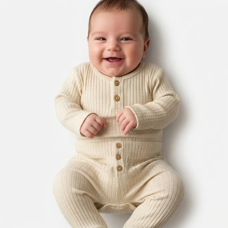 A smiling baby wearing a cream ribbed long-sleeve footless romper with brown buttons, photographed against a clean white background.