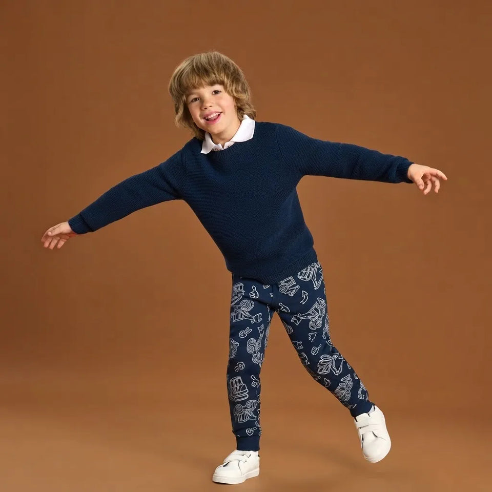 A young boy wearing a navy blue sweater and matching navy patterned pants with white sneakers, posing with outstretched arms against a solid brown studio background.