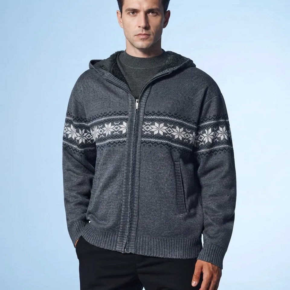 A man wearing a dark gray zip-up knit hooded sweater with a white Nordic snowflake pattern across the chest and sleeves, paired with a dark gray t-shirt and black pants, set against a light blue studio background.