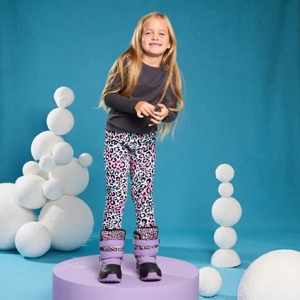 A young girl with long blonde hair stands on a lavender platform wearing patterned leggings with a pink, black, and white leopard print, a dark gray long-sleeve shirt, and purple ski boots. The background is a bright blue studio backdrop decorated with white stacked foam spheres.