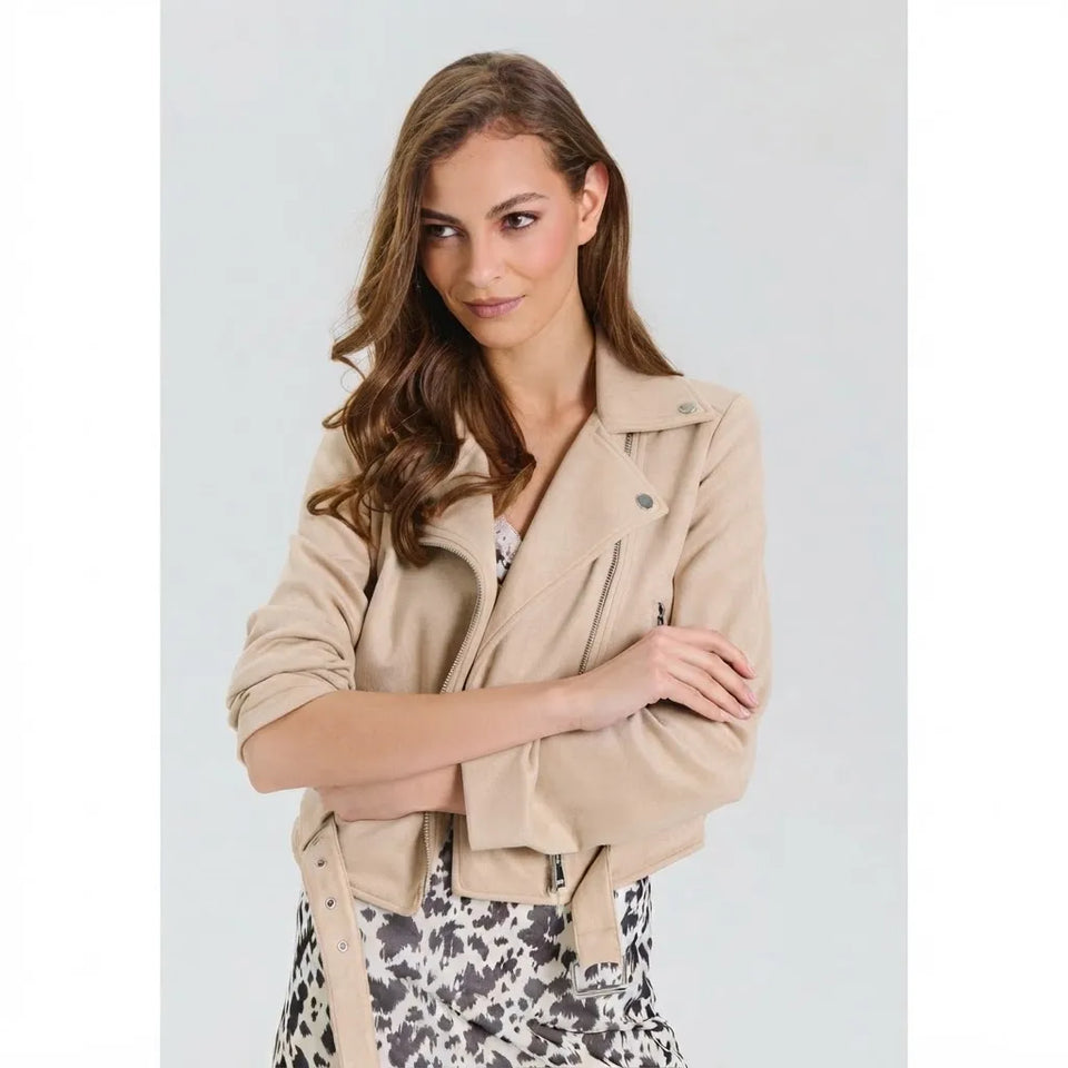 A woman modeling a cropped beige moto jacket with a matching tie belt, paired with a black and white patterned skirt, against a light gray studio background.