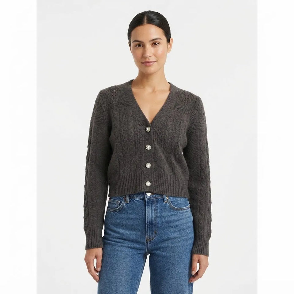A woman modeling a dark brown cable-knit cropped cardigan with a V-neck and silver buttons, paired with blue jeans, against a plain light gray background.