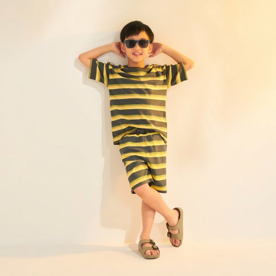 A child wearing a matching two-piece striped lounge set with a short-sleeve top and shorts in olive green and yellow, styled with dark sunglasses and brown sandals against a plain light background.
