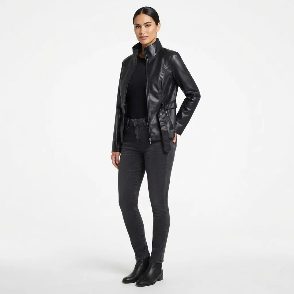 A woman modeling a black faux-leather moto jacket with a high collar, zippered front, and a matching black belt, styled with dark gray skinny jeans and black ankle boots against a plain white studio background.
