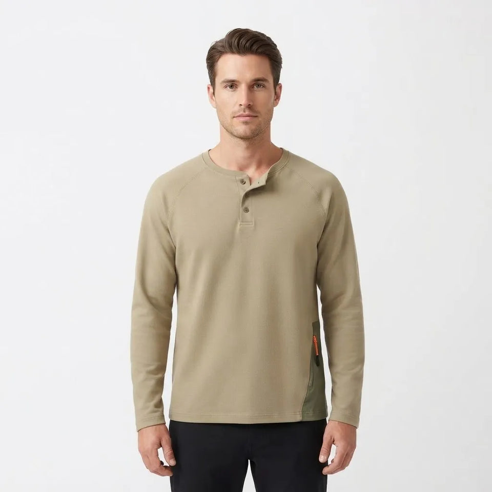 A man wearing a light khaki long-sleeve henley shirt with dark pants, standing against a plain white studio background.