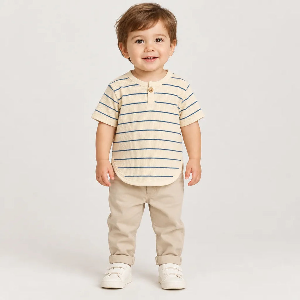 A toddler standing against a light gray studio background, wearing a cream short-sleeve henley shirt with thin blue horizontal stripes, beige pants, and white shoes.