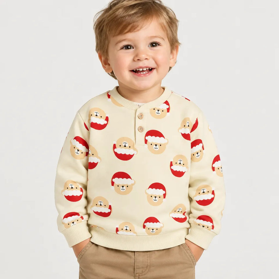 A young child wearing a cream-colored long-sleeve henley sweatshirt with a repeating pattern of Santa Claus faces, paired with khaki pants, against a plain light gray background.