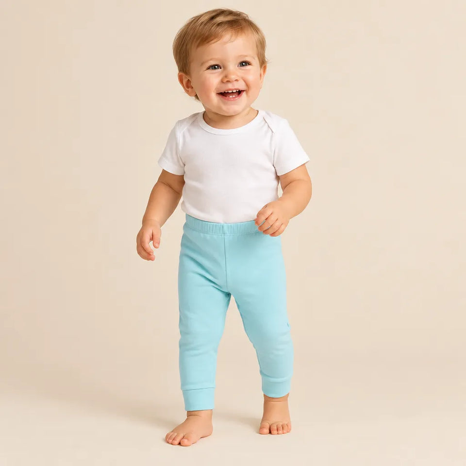 A smiling toddler standing barefoot against a warm beige studio background, wearing a plain white short-sleeve top and light aqua pants.