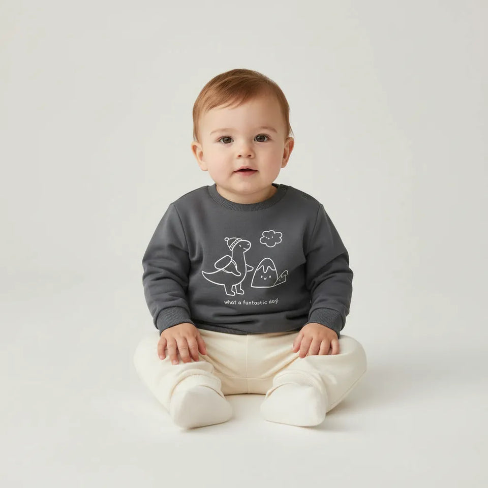 A baby sitting cross-legged on a light gray studio background, wearing a dark gray long-sleeve sweatshirt with a white dinosaur graphic and cream pants.