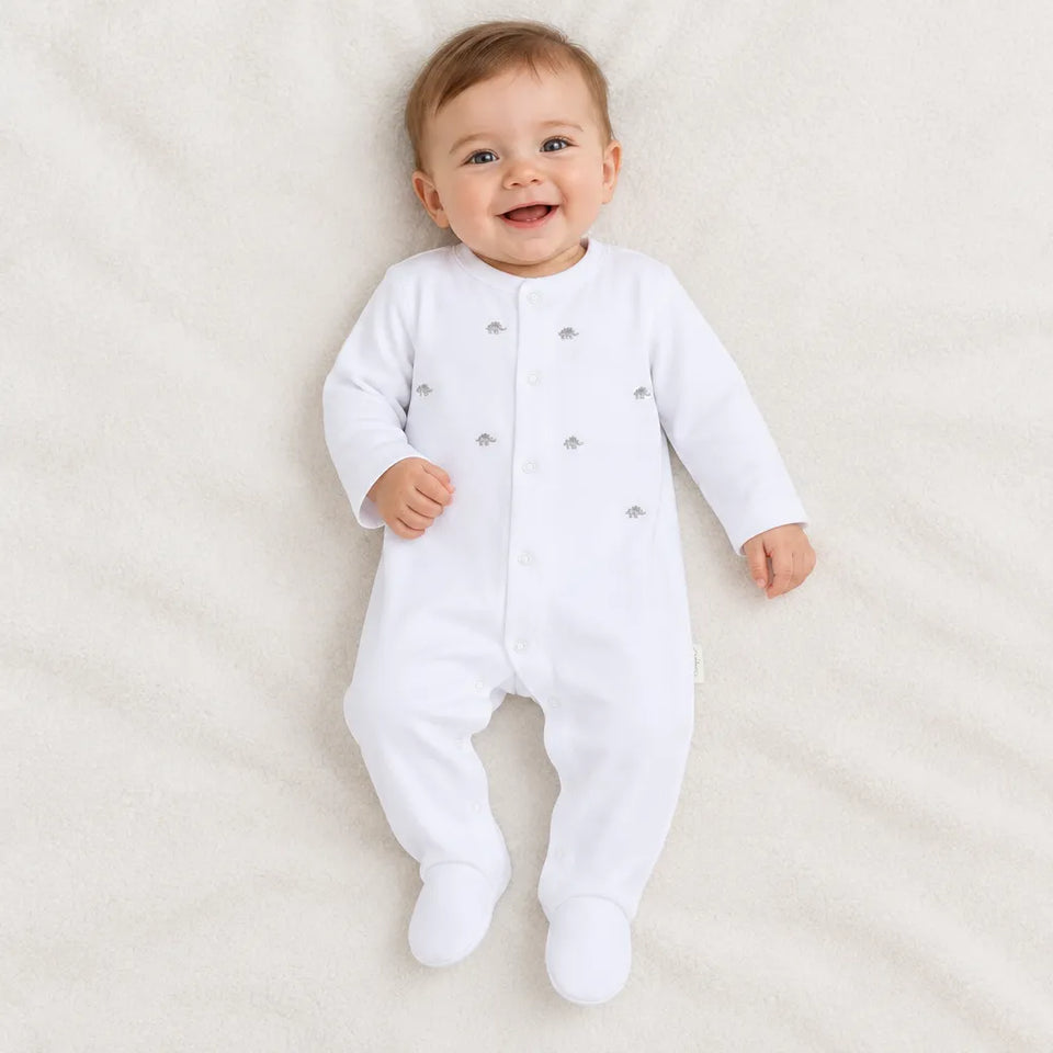A baby lying on a soft cream-colored blanket, wearing a white long-sleeve footed sleeper with small silver decorative accents.