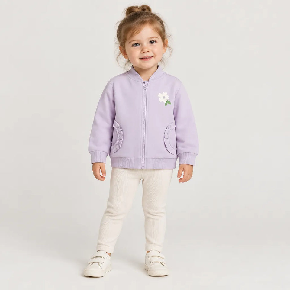 A young child stands against a light gray studio background wearing a lavender zip-front jacket with a small white flower applique on the chest, cream pants, and white shoes.