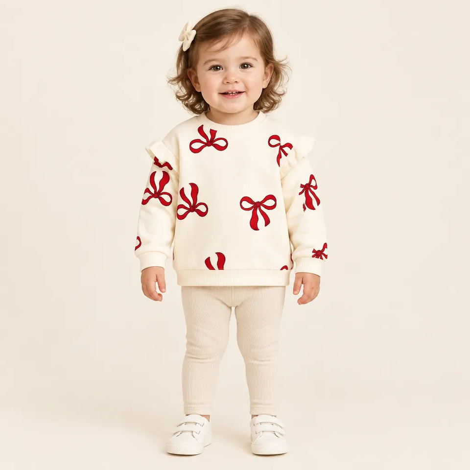A toddler girl stands against a warm off-white studio background wearing a cream sweatshirt with red ribbon patterns, matching cream leggings, and white shoes.
