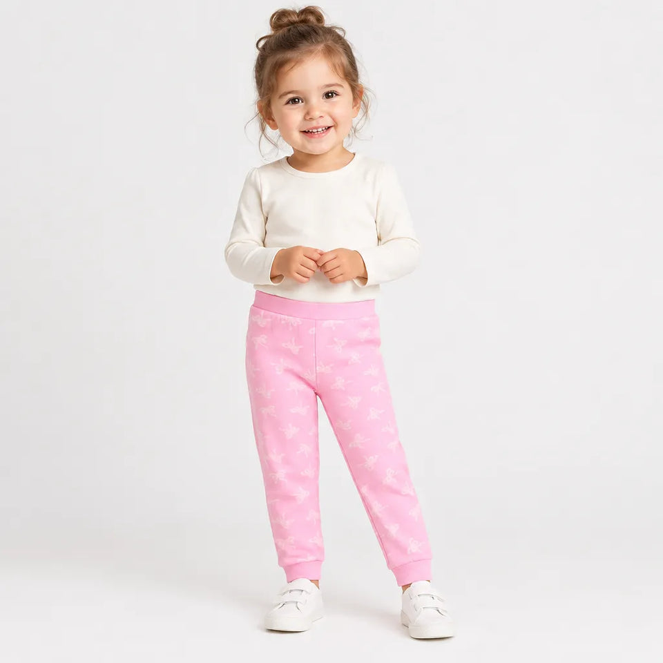 A young child wearing pink patterned sweatpants and a cream long-sleeve top, standing against a plain white studio background.