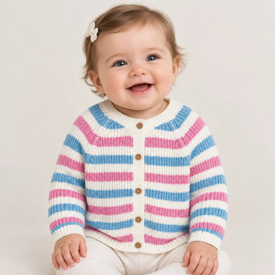A smiling baby wearing a knitted striped cardigan with pink, blue, and white horizontal stripes and brown buttons, paired with cream pants, set against a plain light beige background.