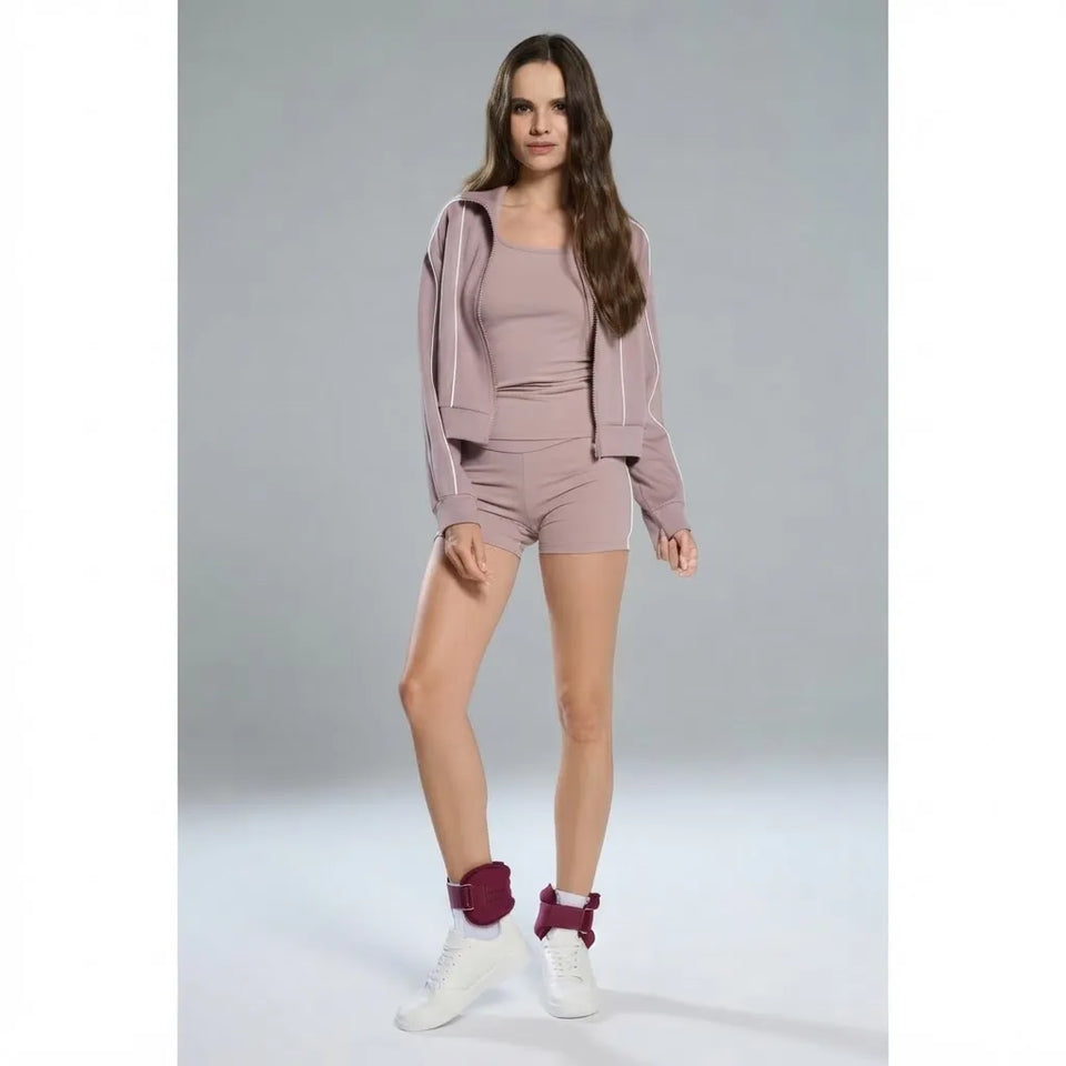A woman modeling a matching mauve workout set consisting of a zip-up hoodie, tank top, and shorts, paired with white sneakers and burgundy ankle cuffs, against a plain gray studio background.