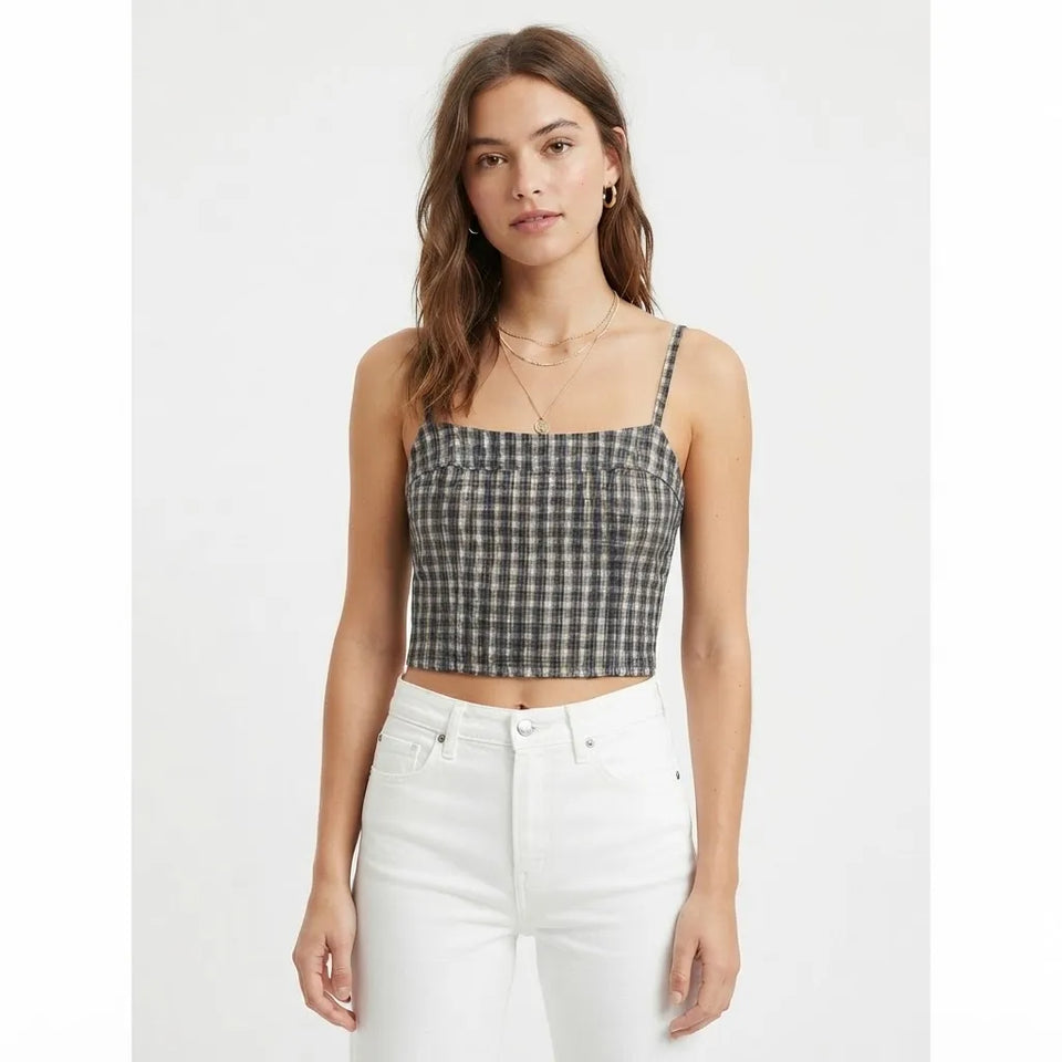 A woman modeling a black and white checkered crop top with thin spaghetti straps, paired with white jeans, against a plain light gray background.