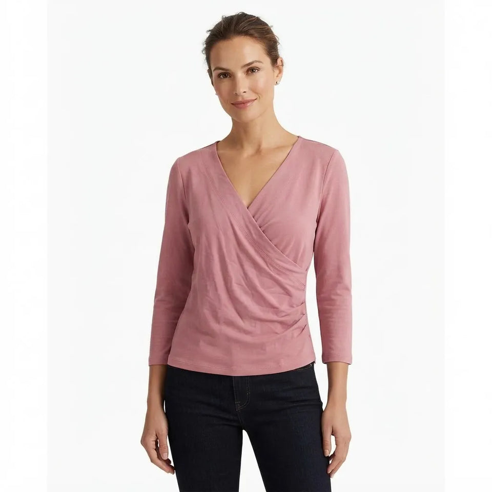 A woman modeling a dusty pink wrap-style long-sleeve top with dark jeans against a plain white studio background.