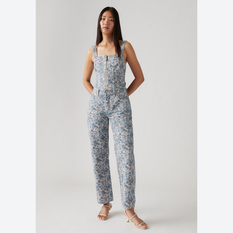 A woman modeling a sleeveless jumpsuit with a blue and beige floral print, standing against a light gray studio background.