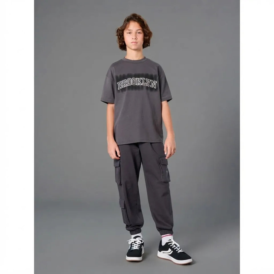 A young boy stands against a solid gray studio background wearing a dark gray crewneck t-shirt with a distressed 'BROOKLYN' graphic across the chest, dark gray cargo jogger pants, and black and white sneakers.