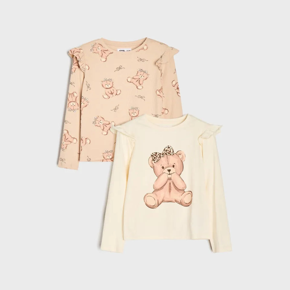 Two children's long-sleeve tops with ruffled shoulders, displayed on a light gray background. The top left is a light peach color with a repeating teddy bear print, and the top right is a cream color with a large central teddy bear graphic wearing a flower crown.