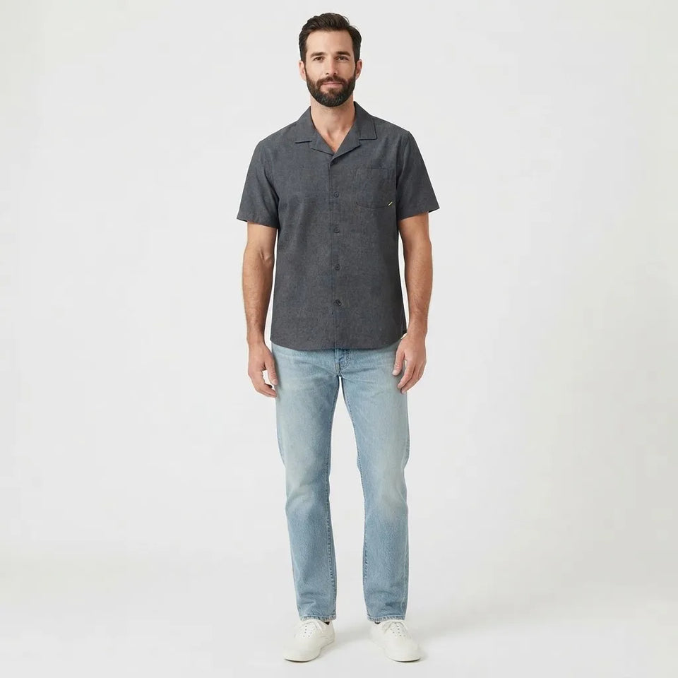 A man wearing a dark gray short-sleeve button-up shirt, light blue jeans, and white sneakers, standing against a plain white studio background.