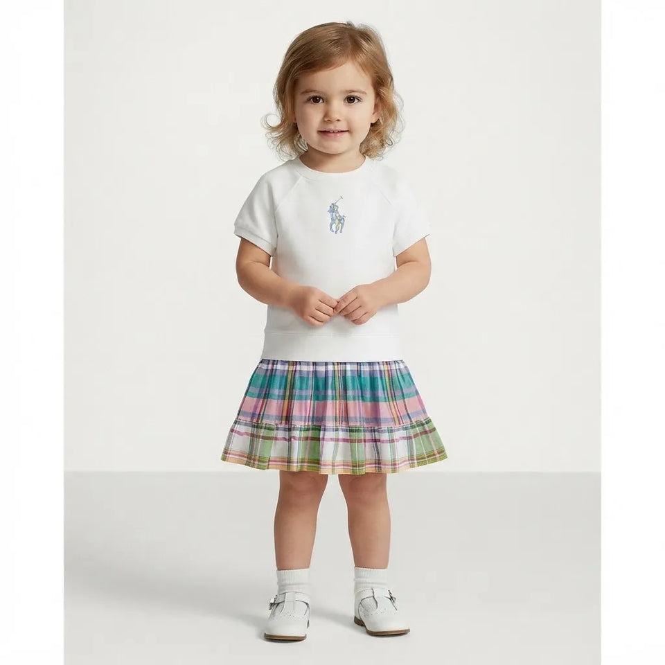 A toddler girl stands against a plain light gray studio background wearing a white short-sleeve top with a small blue Polo pony logo on the chest and a multicolor plaid skirt in pink, blue, green, and yellow tones. She is also wearing white socks and white shoes.