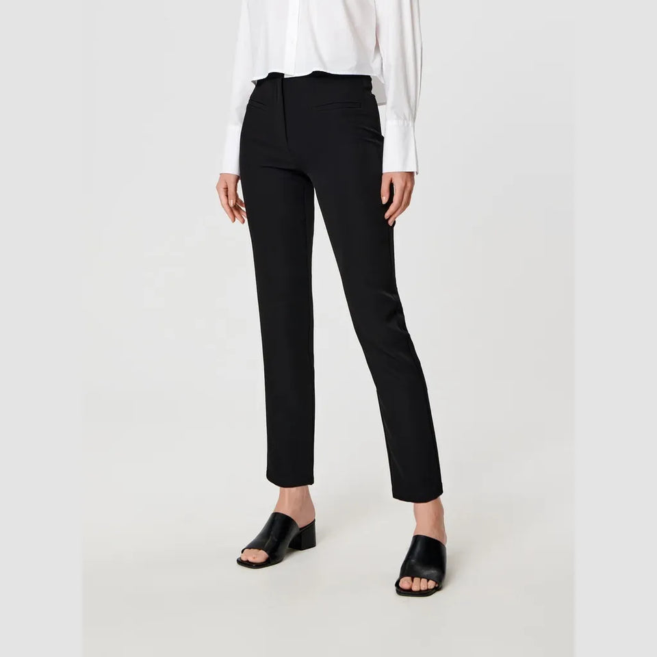 A model wearing black ankle-length straight-leg trousers, styled with a white long-sleeve blouse and black slide sandals, photographed against a light gray studio background.