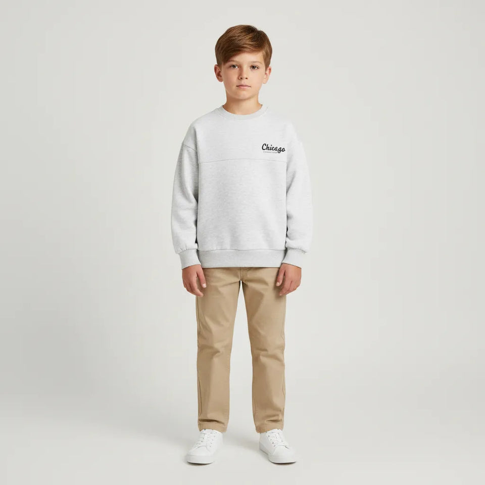 A young boy stands against a light gray studio background wearing a light gray crewneck sweatshirt with a black 'Chicago' logo on the chest, khaki pants, and white sneakers.