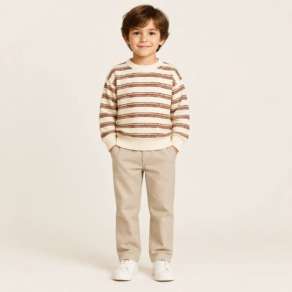 A young boy stands against a plain light beige studio background wearing a cream and brown horizontally striped long-sleeve knit sweater, beige pants, and white sneakers.