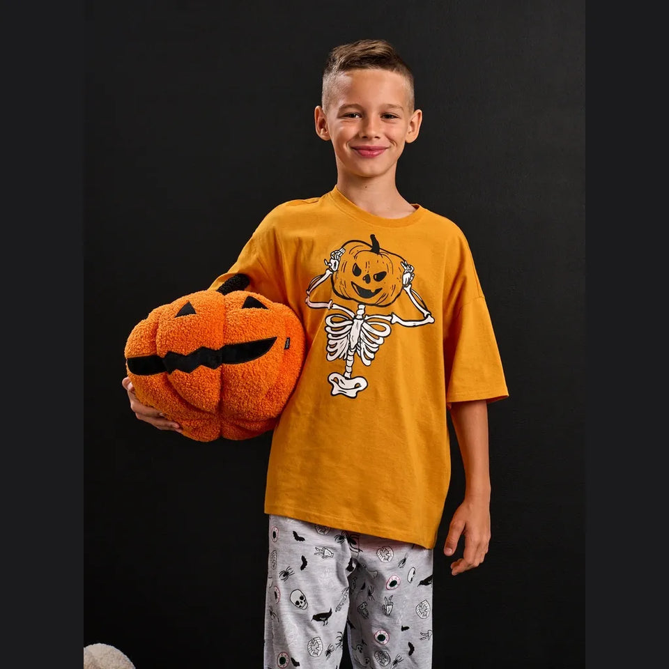 A young boy wearing a yellow-orange t-shirt with a pumpkin skeleton graphic and patterned pajamas, holding a plush jack-o'-lantern against a black background.