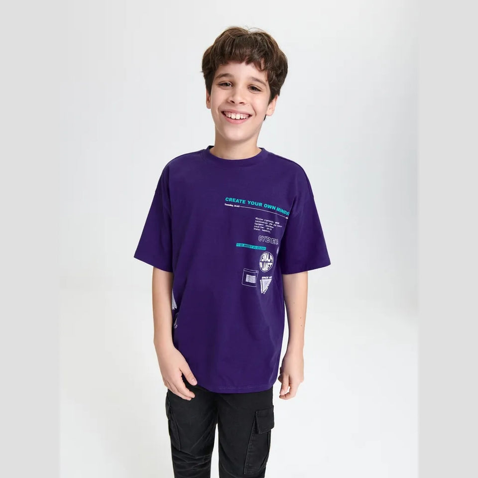 A smiling young boy wearing a purple short-sleeve graphic t-shirt and black cargo pants, standing against a light gray studio background.