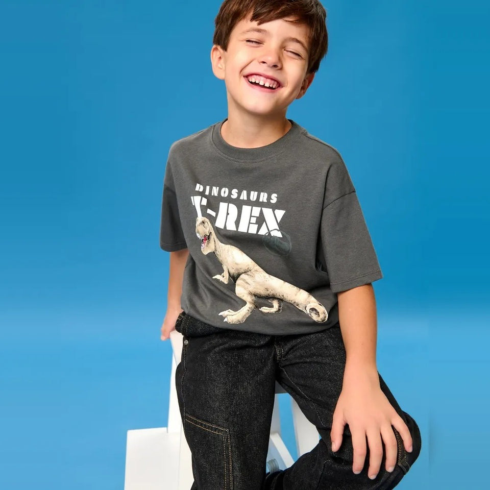 A young boy wearing a dark gray graphic t-shirt with a white 'DINOSAURS T-REX' print and dark jeans, sitting on a white stool against a solid blue background.