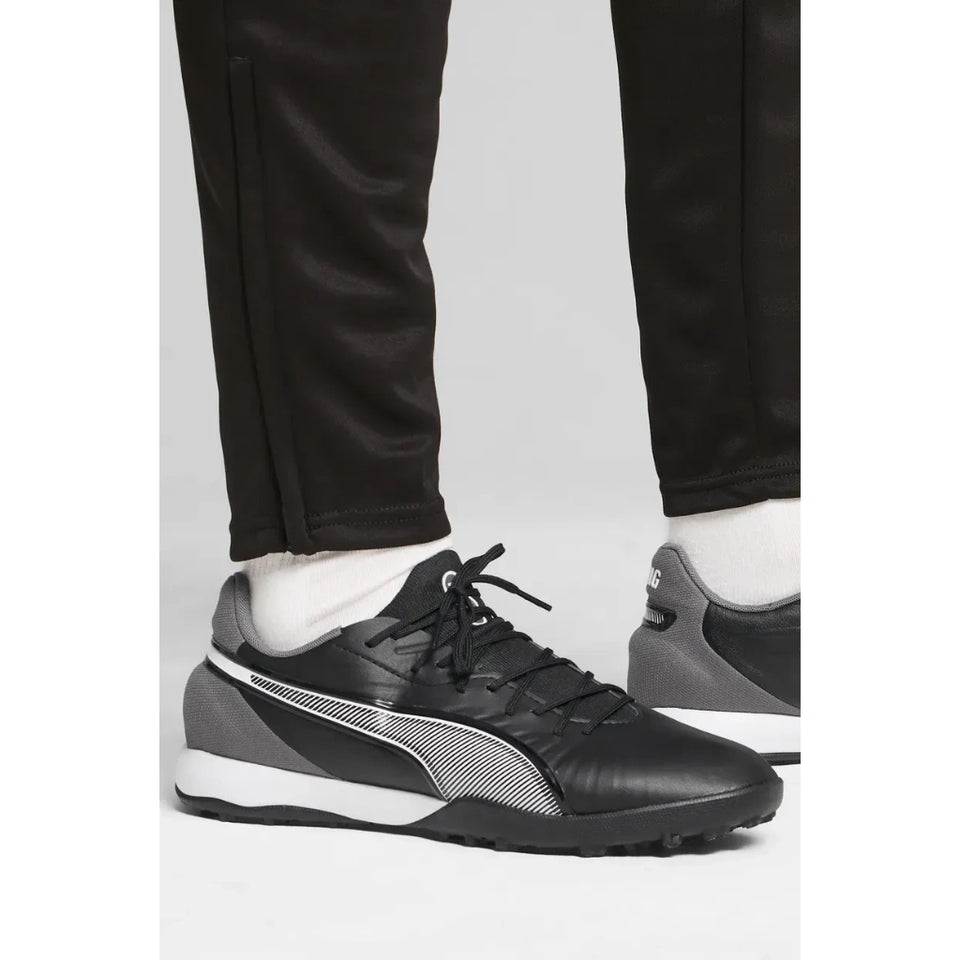 Black Puma turf shoes with white and gray accents, shown on a model wearing black pants and white socks against a light gray background.