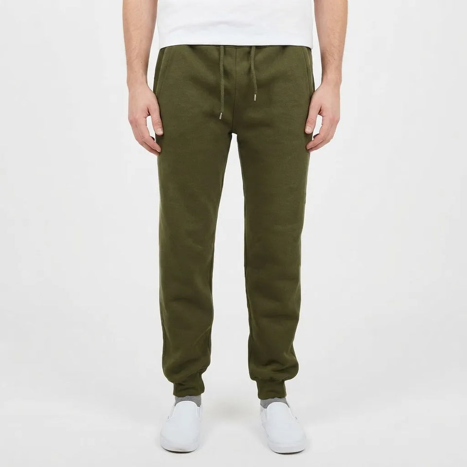 A person wearing olive green jogger sweatpants with a drawstring waist, paired with a white t-shirt and white slip-on shoes, shown from the waist down on a light gray studio background.