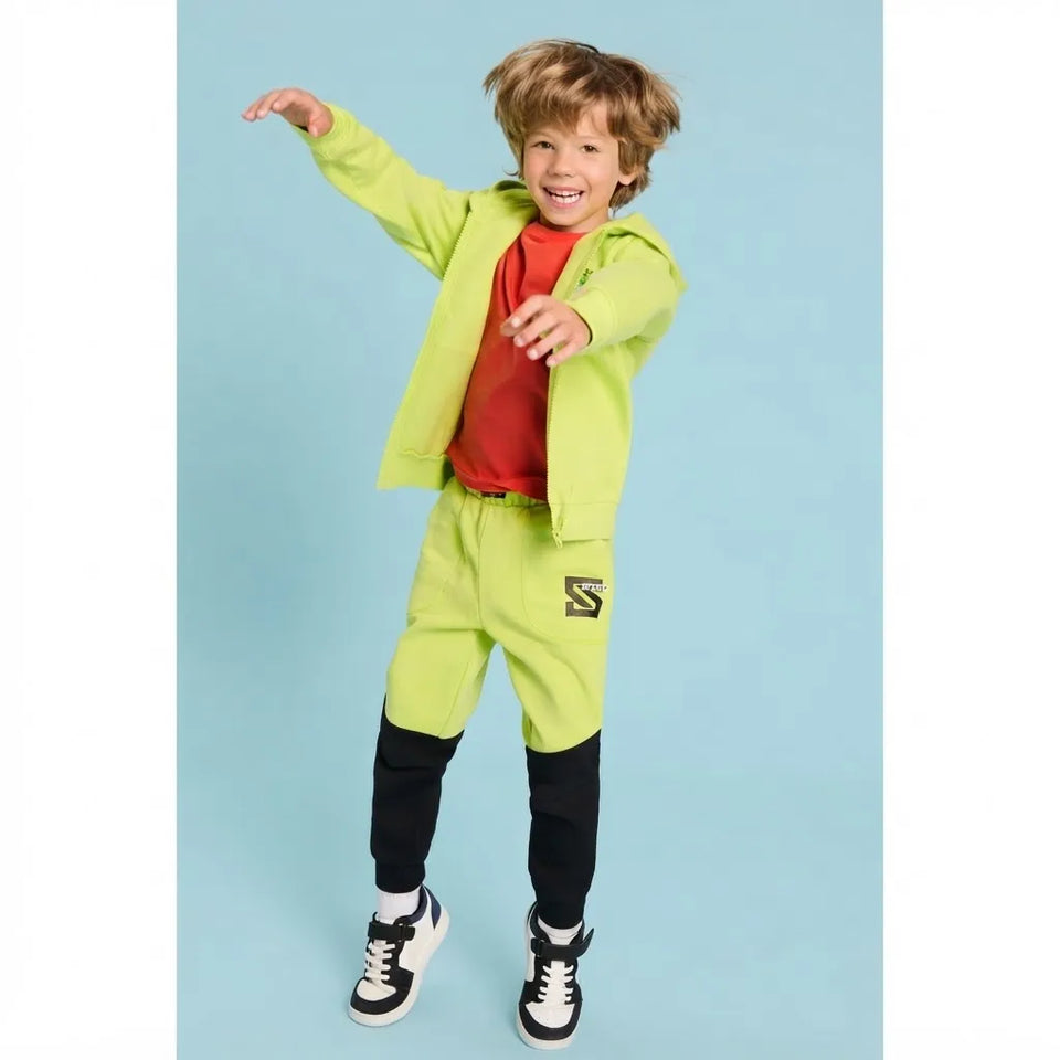 A young boy modeling a bright lime green tracksuit consisting of a zip-up hoodie, matching sweatpants, and black knee-high socks, paired with a red t-shirt and black-and-white sneakers, set against a solid light blue background.