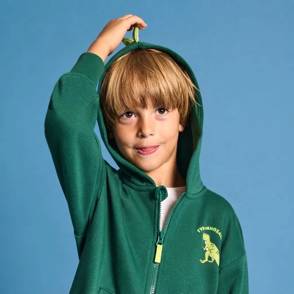 A young boy with blonde hair wearing a green zip-up hoodie with a dinosaur graphic on the chest, posing against a solid blue background.