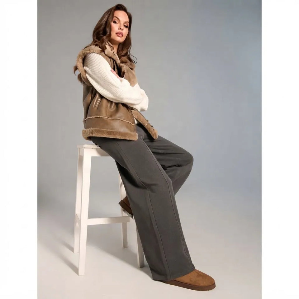 A woman sits on a white stool wearing a brown shearling vest over a cream knit sweater, dark gray wide-leg pants, and brown suede boots against a light gray studio background.