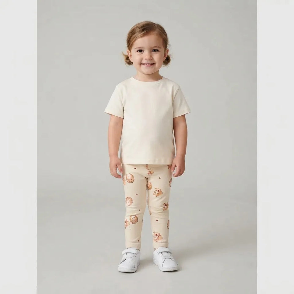 A toddler girl stands against a light gray studio background wearing a cream short-sleeve t-shirt and matching cream leggings with a small animal print, paired with white sneakers.