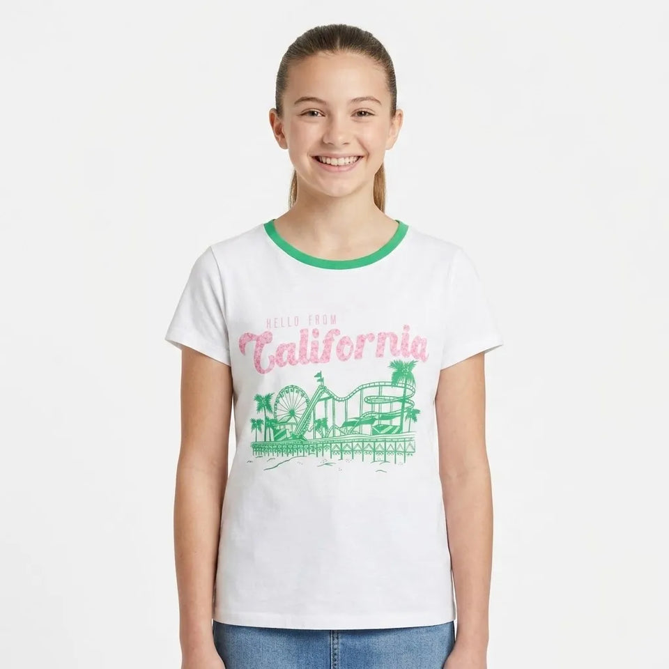 A young girl smiling and wearing a white ringer t-shirt with a green crew neck and a pink and green 'Hello from California' graphic on the front, paired with blue jeans, against a plain white background.