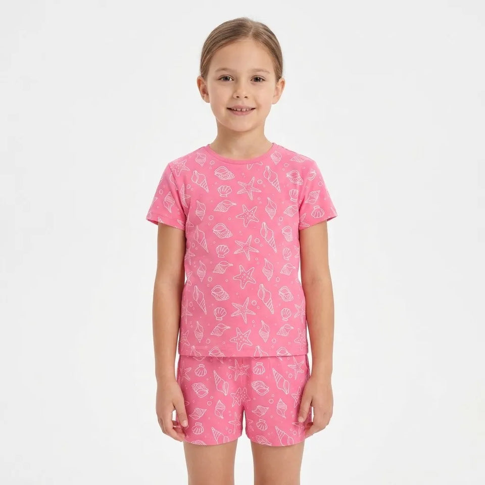 A young girl modeling a pink short-sleeve t-shirt and matching shorts set with a white seashell pattern, set against a plain white studio background.