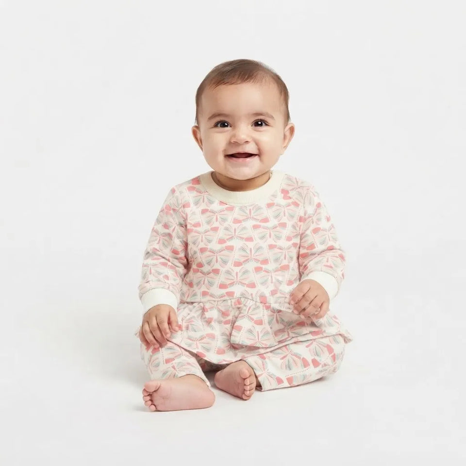 A smiling baby sitting on a white studio background, wearing a long-sleeve romper pajama with a pink and gray butterfly pattern on a cream base.