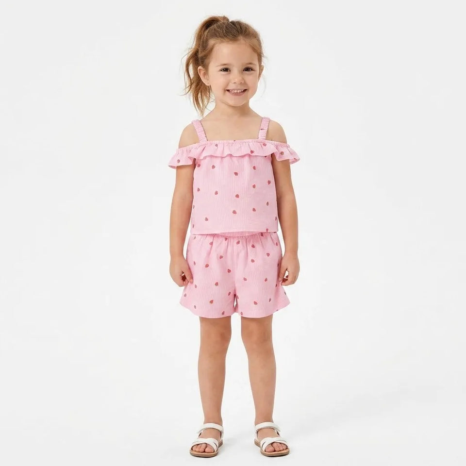 A young girl with her hair in a ponytail stands against a plain white studio background wearing a pink two-piece outfit with small red polka dots. The set includes a cold-shoulder top with ruffled trim and matching shorts.