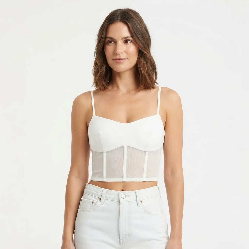 A woman modeling a white spaghetti-strap crop top with a sheer mesh corset-style panel, paired with white jeans, against a plain white studio background.
