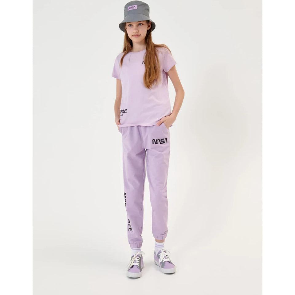 A young girl modeling a light purple/lavender matching set consisting of a short-sleeve t-shirt and jogger sweatpants, styled with a gray bucket hat, white socks, and purple sneakers against a plain white studio background.