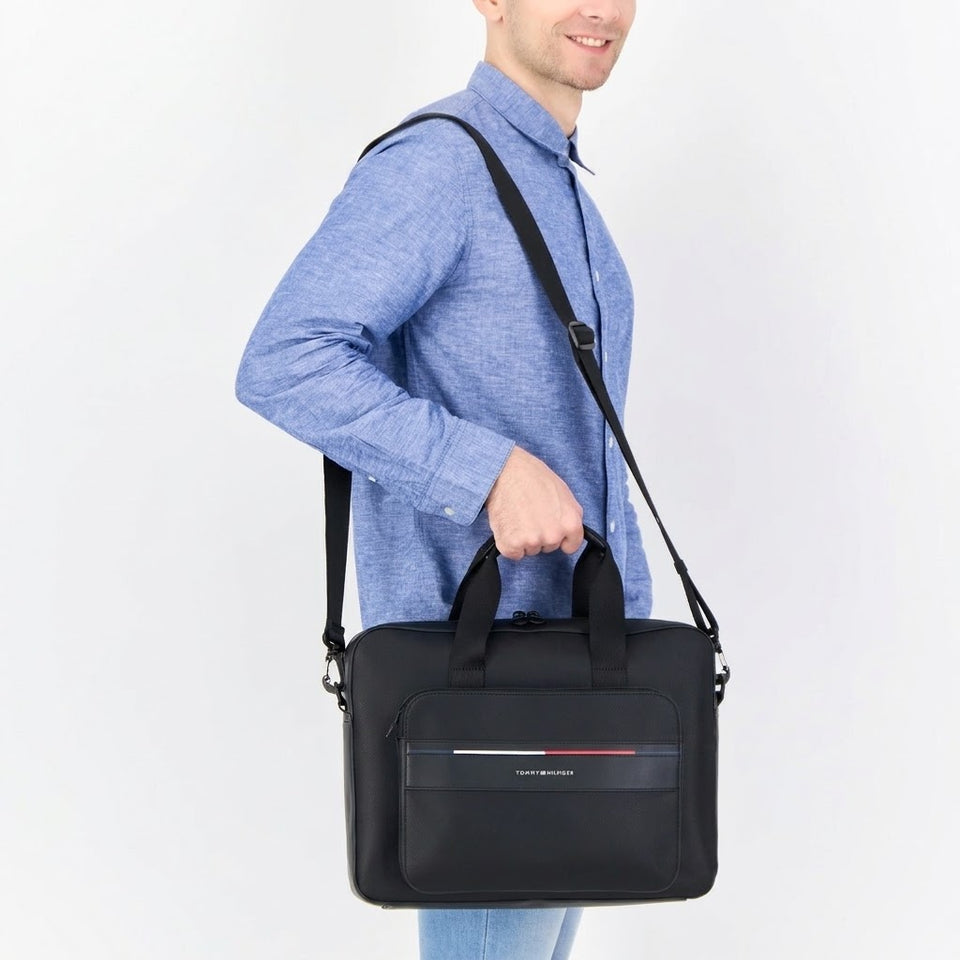 A man in a blue shirt carries a black briefcase-style shoulder bag with a top handle, front zip pocket, and a small red and white stripe detail on the front, shown against a plain white background.