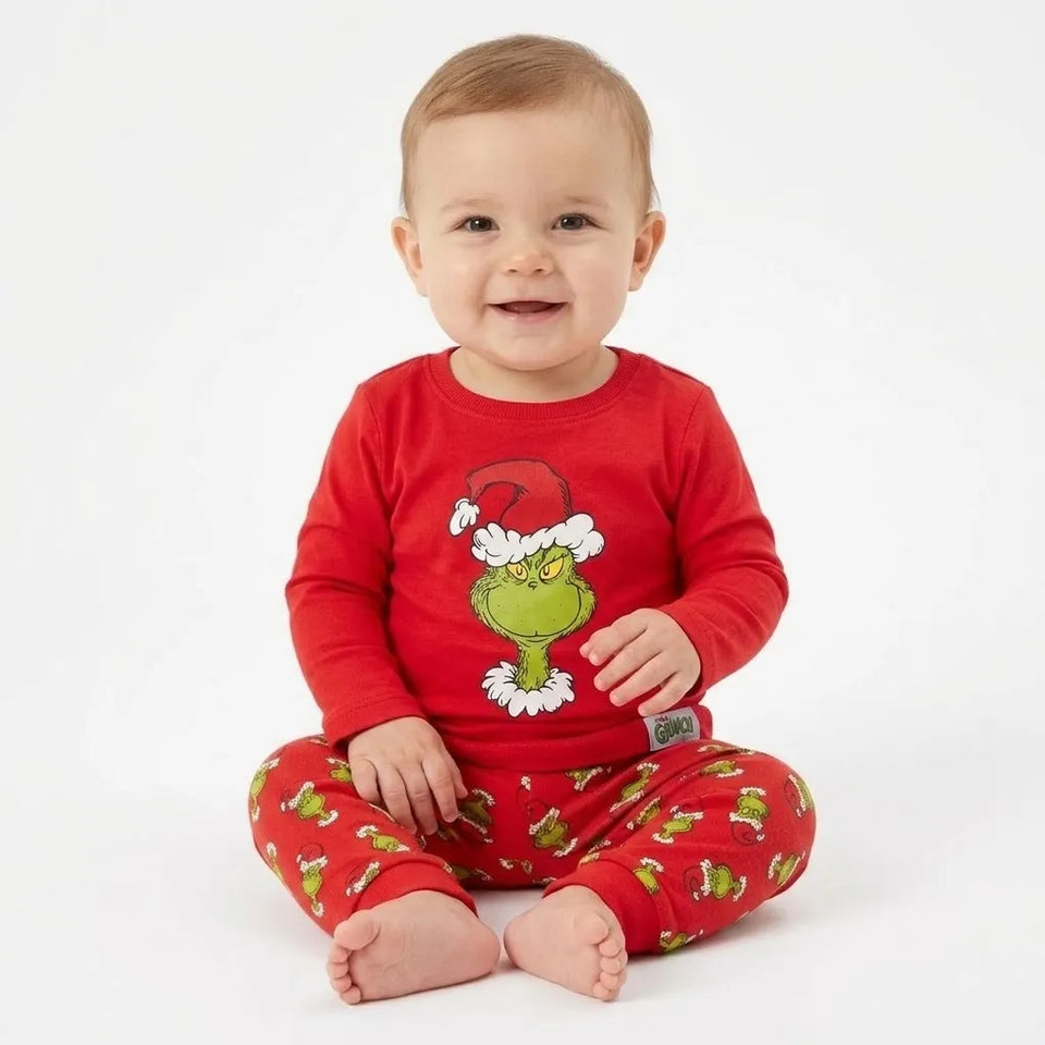 A baby sitting cross-legged wearing a red long-sleeve Grinch pajama top and matching red pajama pants with a green Grinch print, against a plain white background.