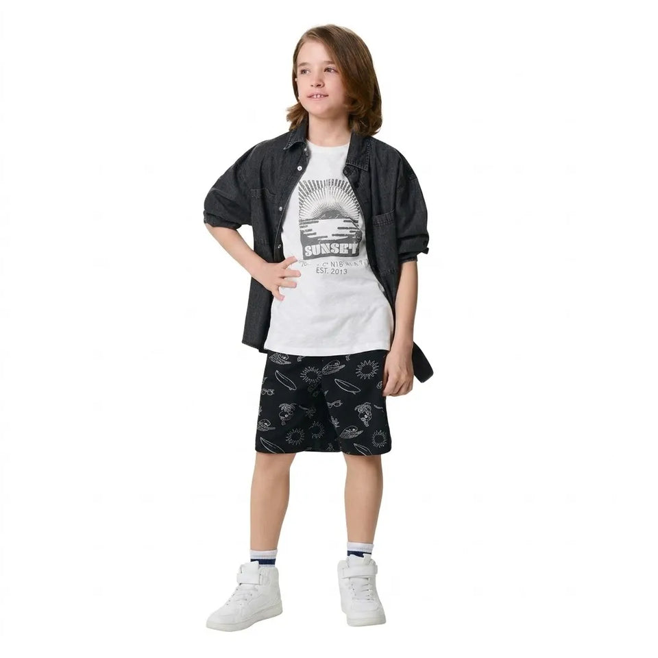 A child model wearing a casual outfit consisting of a white graphic t-shirt, a black button-up shirt, black patterned shorts, white socks, and white sneakers, set against a plain white background.