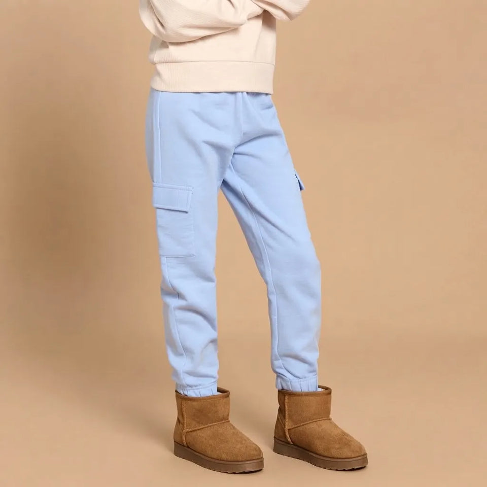 A child wearing light blue cargo jogger pants with elastic cuffs, styled with a cream sweatshirt and brown suede ankle boots against a solid beige studio background.