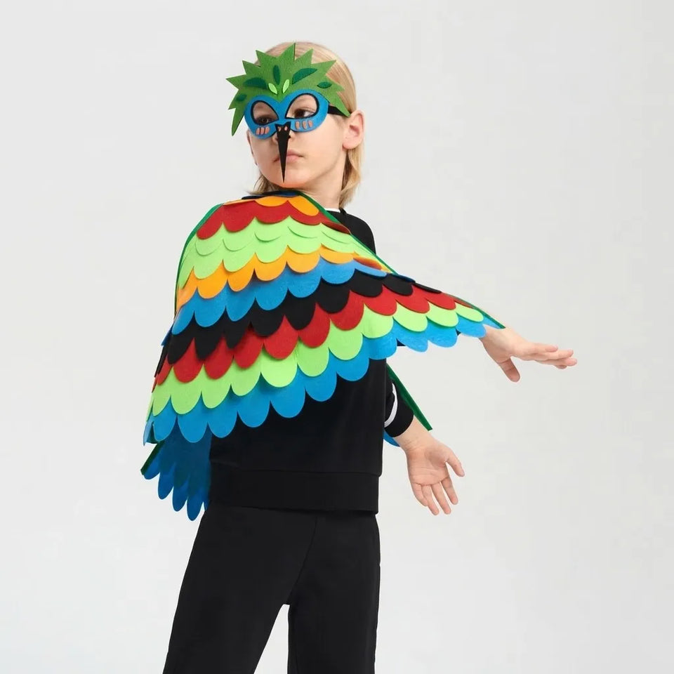 A child wearing a colorful bird costume cape with layered green, yellow, orange, red, black, and blue feathers, along with a matching green and blue mask, posed against a light gray background.